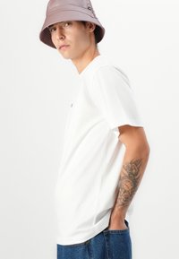 Weißes Baumwoll-T-Shirt mit lockerer Passform, kurzen Ärmeln und einem kleinen blauen Logo auf der Brust. Kombiniert mit Jeans-Shorts. Helllila Bucket Hat.