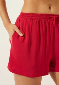 Červené šortky vyrobené z lehkého materiálu s elastickým pasem, detaily na šňůrku a kapsami po obou stranách, hladká textura.