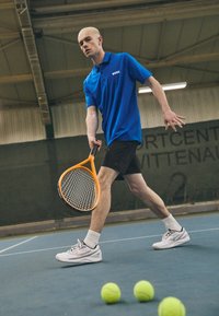 Blå polo t-shirt med "BOSS" logo, sorte shorts, hvide tennissko, holder en orange tennisketsjer. Tre gule tennisbolde på banen.