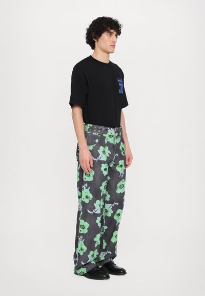 Schwarzes T-Shirt mit blauem Grafikdruck, kombiniert mit lässigen grauen Jeans in lockerem Schnitt, die ein großes grünes Blumenmuster haben, sowie einem grünen Nieten-Gürtel.