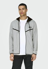 Sudadera gris con cremallera y capucha forrada de marrón, que presenta detalles en negro y texturizados. El modelo lleva una camiseta blanca debajo.
