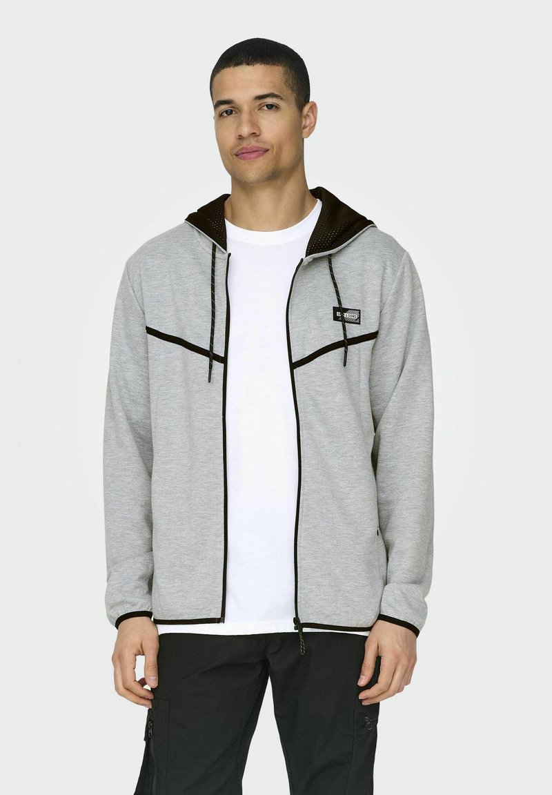Sudadera gris con cremallera y capucha forrada de marrón, que presenta detalles en negro y texturizados. El modelo lleva una camiseta blanca debajo.