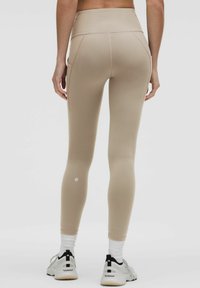 Leggings beige a vita alta con una texture liscia e elastica e un design aderente. Presentano tasche laterali e un logo sulla parte inferiore della gamba.