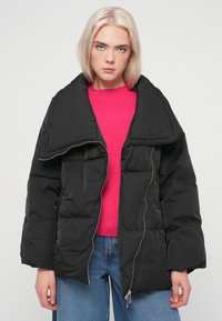 Μαύρο μπουφάν με επένδυση, ευρύ γιακά, κλείσιμο με φερμουάρ και μεγάλες τσέπες, φορεμένο πάνω από ένα φωτεινό ροζ πουλόβερ, συνδυασμένο με μπλε τζιν.