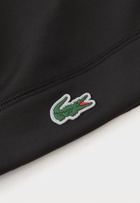 Črn material z zelenim logotipom krokodila in rdečimi detajli, prišit na spodnjem robu. Struktura površine kaže rahlo sijaj.