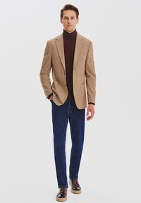 Brązowa marynarka w kratę noszona na ciemnym golfie, w zestawieniu z ciemnoniebieskimi dżinsami i brązowymi trampkami z jasną podeszwą. Prosty, dopasowany fason.