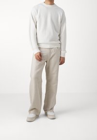Uomo che indossa un maglione bianco a maniche lunghe con texture, pantaloni beige a gamba larga e sneakers bianche, in piedi contro uno sfondo bianco uniforme.
