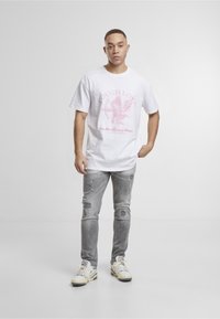 Jeune homme debout, une main dans la poche, portant un t-shirt graphique blanc "Tough Love", un jean gris déchiré et des baskets blanches sur fond uni.