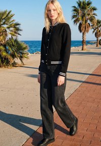 Young woman wearing black varsity jacket, dark jeans, and black shoes walking along a seaside path lined with palm trees.