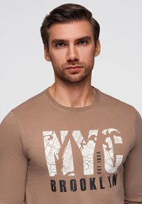 Langärmliges, braunes Baumwollhemd mit auffälligem "NYC" und "Brooklyn"-Text in Weiß und Schwarz sowie Kartendetails. Glatte Textur, taillierter Schnitt.