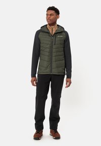 Giacca da uomo imbottita di colore verde scuro con texture trapuntata, maniche nere, cappuccio e tasca con zip. Abbinata a pantaloni neri e stivali marroni.