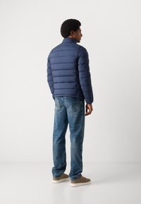 Doudoune marine avec un design matelassé horizontal, col haut et texture lisse, portée avec un jean bleu et des baskets beiges.
