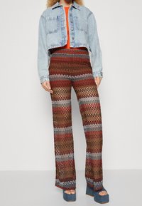 Denimkort jacka, orange topp, mönstrade utsvängda byxor med röda, bruna och silverränder. Blå plattformsskor med öppen tå.