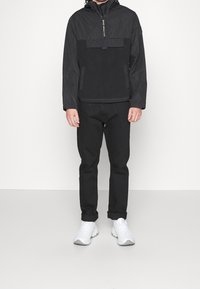 Chaqueta negra con cierre frontal, capucha y bolsillo en el pecho; combinada con pantalones negros y zapatillas blancas. Textura suave y diseño moderno.
