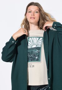 Camicia button-up verde scuro con un motivo testurizzato, indossata sopra una t-shirt beige con una grafica che presenta un design di sole e acqua.