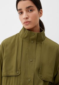 Junge Frau mit dunklem Haar, das zurückgebunden ist, trägt eine olivgrüne Jacke mit hohem Kragen und Druckknöpfen vor einem schlichten hellen Hintergrund.