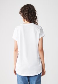 Camiseta de algodón blanca con mangas cortas, escote redondeado y un dobladillo trasero más largo, combinada con jeans de mezclilla azul. Textura suave, sin patrones.