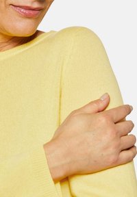 Helles gelbes Sweatshirt mit gerippter Textur und weitem Ausschnitt. Eine Hand ruht auf der Schulter und hebt das weiche Material des Kleidungsstücks hervor.