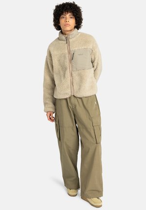 Beige fleece jack met hoge kraag en een voorkennis; gecombineerd met olijfgroene cargobroek met meerdere zakken; bruine schoenen.