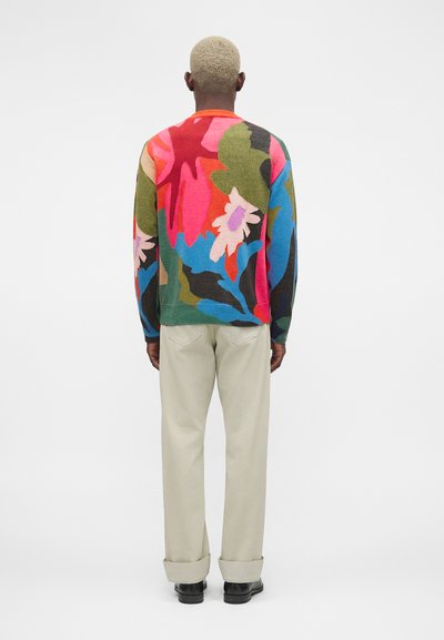 Άνδρας με κοντά ξανθά μαλλιά που φοράει πολύχρωμο αφηρημένο φλοράλ πουλόβερ, μπεζ παντελόνι και μαύρα παπούτσια, στέκεται γυρισμένος με την πλάτη.