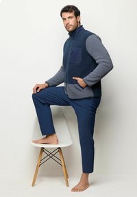 Fleece-vesti tummansinisenä harmailla yksityiskohdilla, jossa on vetoketjullinen tasku, käytetään sinisten housujen päällä; malli seisoo yhdellä jalalla tuolilla, paljain jaloin.