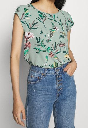 Femme portant un chemisier vert à manches courtes avec imprimé floral, rentré dans un jean bleu taille haute à multiples boutons sur le devant, la main dans la poche.