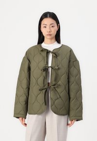 Oliven grønn quilted jakke med rund krage, dekorert med tre sløyfefestinger og lange ermer, iført over en lys topp og bukser.