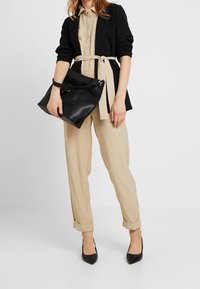 Kobieta ubrana w beżowy kombinezon z paskiem, czarną marynarkę i czarne szpilki z ostrym noskiem, trzymająca dużą czarną skórzaną torebkę na neutralnym tle.