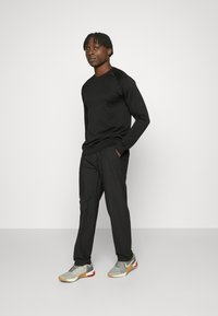 Svart strukturerad långärmad topp kombinerad med löst sittande svarta byxor och grå sneakers med röda accenter, mot en helt vit bakgrund.