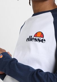 Bílý bavlněný tričko s námořnickými raglánovými rukávy, s barevným logem ellesse na hrudi a detailem loga na rukávu.