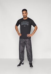 Μαύρο t-shirt Adidas με έντονο λογότυπο, σε συνδυασμό με φαρδιά σκούρα παντελόνια με σχέδια. Μαύρα αθλητικά παπούτσια ολοκληρώνουν το ντύσιμο.