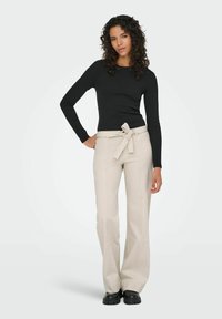 Femme aux cheveux bouclés portant un haut noir à manches longues, un pantalon beige taille haute avec une ceinture nouée, et des chaussures noires épaisses, debout les mains détendues.