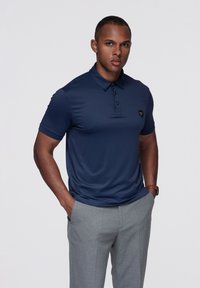Granatowa koszulka polo z krótkim rękawem o gładkiej fakturze i kołnierzykiem, z małym logo na piersi; w połączeniu z jasno szarymi, dopasowanymi spodniami.