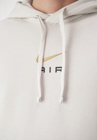 Ljusbeige hoodie med dragsko, prydd med en gyllene Nike-swoosh-logotyp och svart "AIR"-text på framsidan. Mjuk tygtextur.