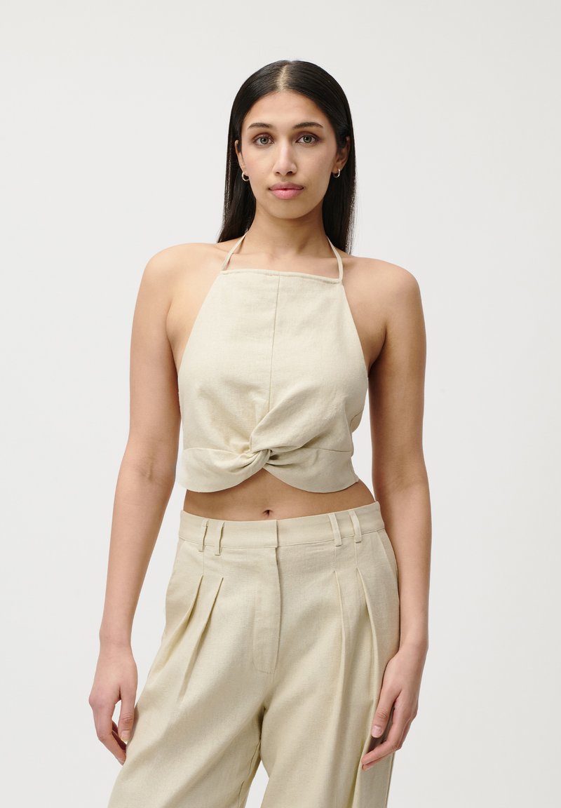 Top cropped beige à col halter avec un détail en nœud à l'avant, associé à un pantalon plissé taille haute assorti, fait d'un tissu léger.