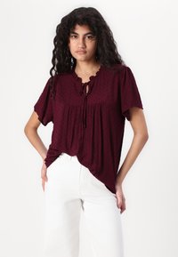 Blusa bordeaux con maniche corte, decorata con un colletto a volant e dettaglio in lacci, caratterizzata da un motivo testurizzato. Abbinata a jeans bianchi.