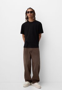 Black cotton t-shirt with a relaxed fit, paired with loose brown denim pants and cream sneakers. Model wearing dark rectangular sunglasses.