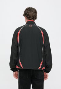 Čierna vetrovka s červenými a bielymi akcentmi, s vysokým golierom, úzkym strihom a elastickými manžetami; výrazné logo na chrbte.