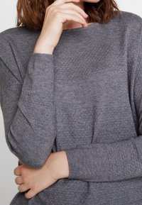 Pull en gris texturé avec un col rond et des manches longues. Le tissu présente un subtil motif en nid d'abeille et des poignets côtelés.