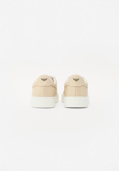 Beige sneakers with a smooth leather exterior, white rubber soles, and a subtle logo embossed on the back. Minimalist design and clean lines.