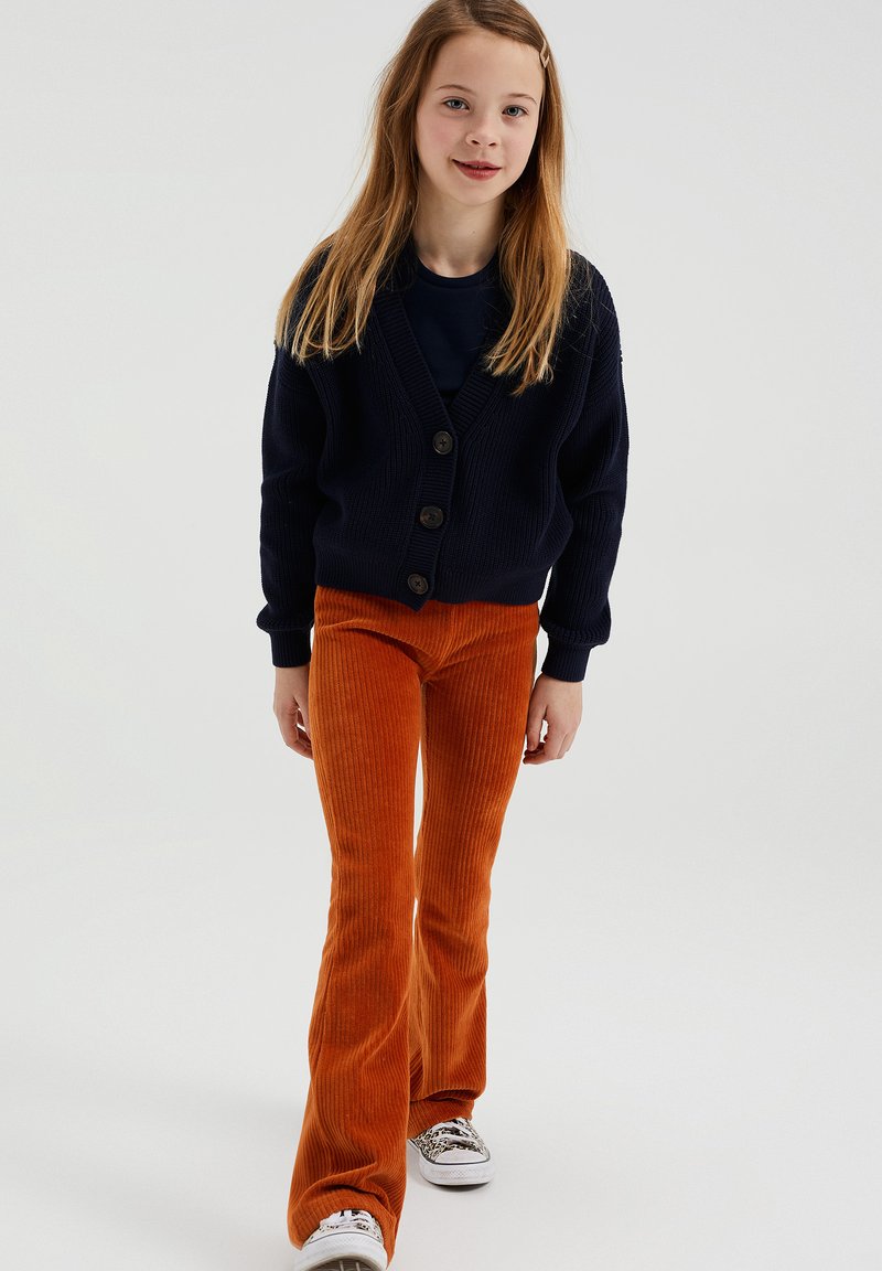 Mornarsko pleteno jopico s petimi gumbi, v kombinaciji z oranžnimi kordnimi širokimi hlačami. Bele teniske s pisanem vzorcem.