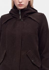 Femme portant une veste en velours côtelé marron foncé avec un col montant, une fermeture éclair, des boutons-pression et des poches à rabat, montrée des épaules jusqu'au milieu du buste.