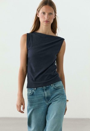 Femme aux longs cheveux portant un haut sans manches bleu marine foncé et un jean taille haute bleu clair, debout devant un fond clair uni.