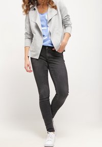 Helle graue Jacke mit drapierter Vorderseite, blaues Hemd mit Grafikdruck, dunkle Skinny-Jeans und weiße Turnschuhe. Lockiges Haar umrahmt das Gesicht.