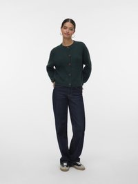Cardigan verde escuro canelado com cinco botões castanhos, combinado com jeans azul escuro e ténis pretos com detalhes cinzentos e brancos.