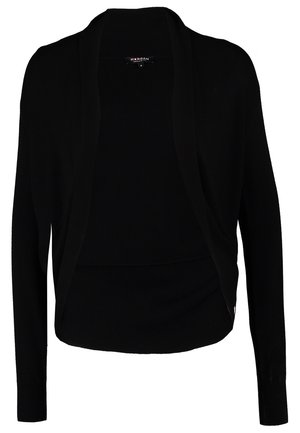 Frau trägt schwarzen Cardigan, weißes Top, dunkle Skinny-Jeans, schwarze Stiefeletten und hält eine schwarze Clutch vor einfarbigem Hintergrund.
