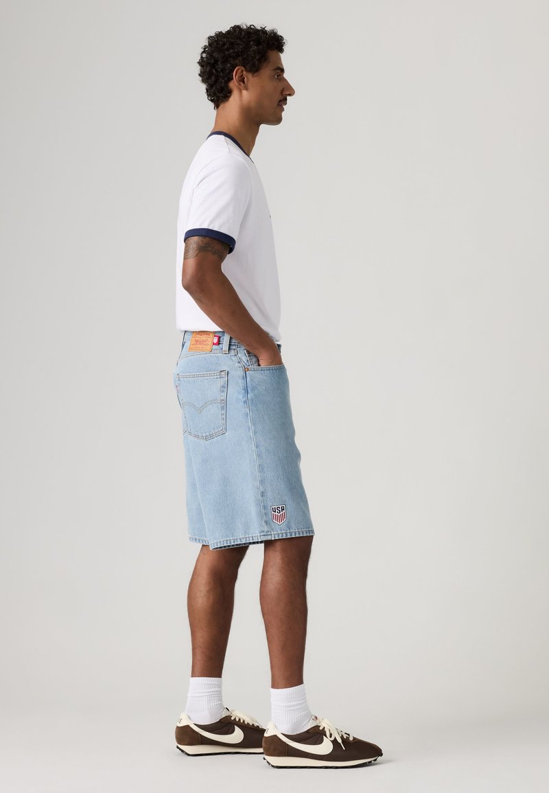Mann trägt weißes T-Shirt und hellblaue Denim-Shorts mit USA-Aufnäher, steht im Profil mit Händen in den Taschen vor einfachem Hintergrund.