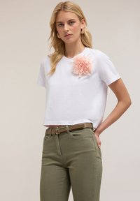 T-shirt bianco a maniche corte con un'applicazione floreale rosa sulla spalla sinistra, abbinato a pantaloni verdi oliva aderenti e una cintura marrone.