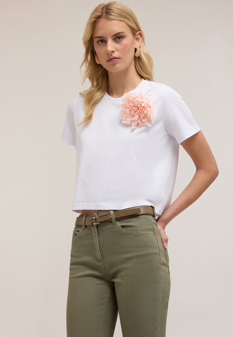T-shirt bianco a maniche corte con un'applicazione floreale rosa sulla spalla sinistra, abbinato a pantaloni verdi oliva aderenti e una cintura marrone.
