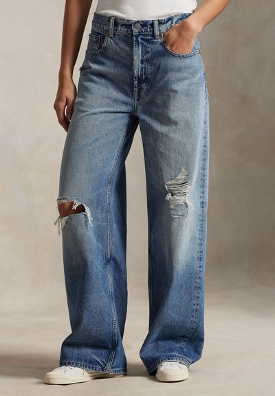 Jeans azzurri a gamba larga con strappi sfilacciati sulle ginocchia, vita alta, classico design a cinque tasche e cuciture a contrasto. Indossati con sneakers bianche.